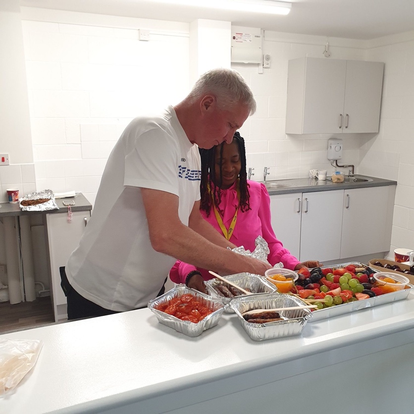 A person and person standing next to a tray of foodDescription automatically generated